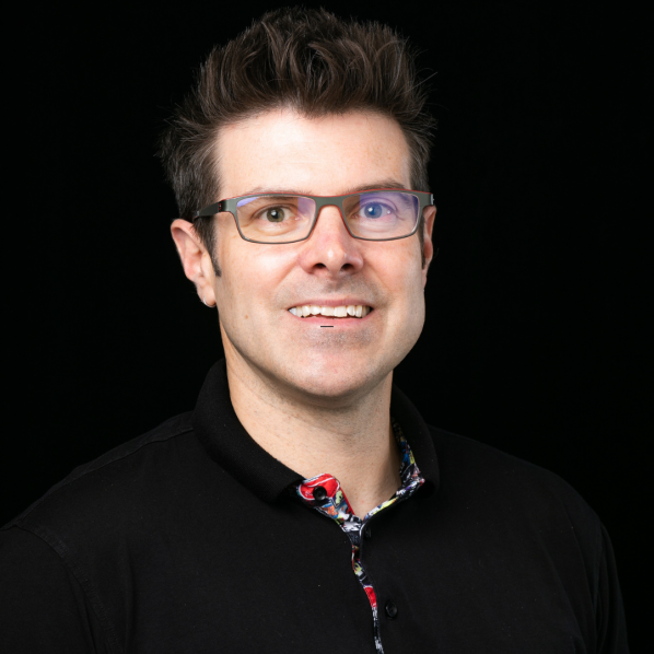 Professional headshot of a smiling man with glasses and dark hair, wearing a black collared shirt against a black background.